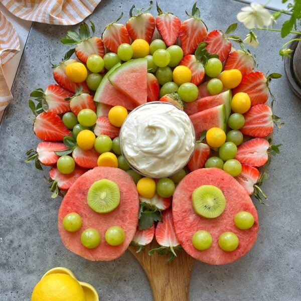 Fruit display with watermelon, strawberries, grapes, mango balls, and kiwi slices are arranged on a wooden board to resemble a bunny.