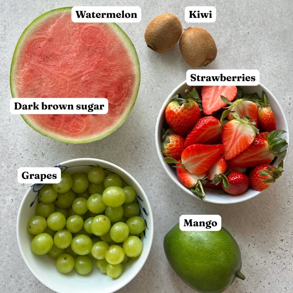 A halved watermelon, two kiwis, a bowl of sliced strawberries, a bowl of green grapes, and a whole mango are displayed on a light surface. Labels identify each fruit.