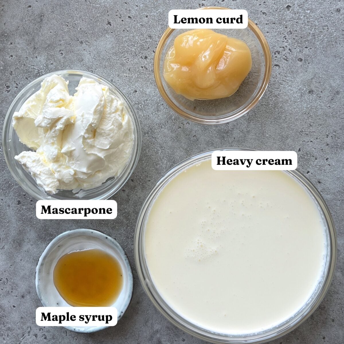 Four labeled bowls on a gray surface, containing mascarpone, maple syrup, lemon curd, and heavy cream.