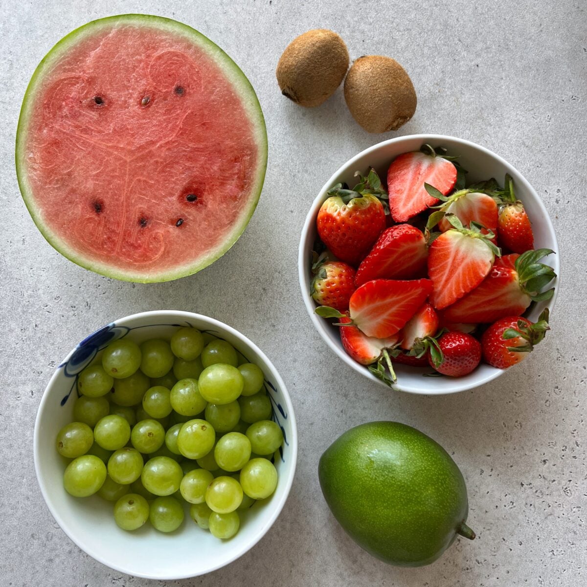 A halved watermelon, two whole kiwis, a bowl of green grapes, a bowl of halved strawberries, and a green mango.