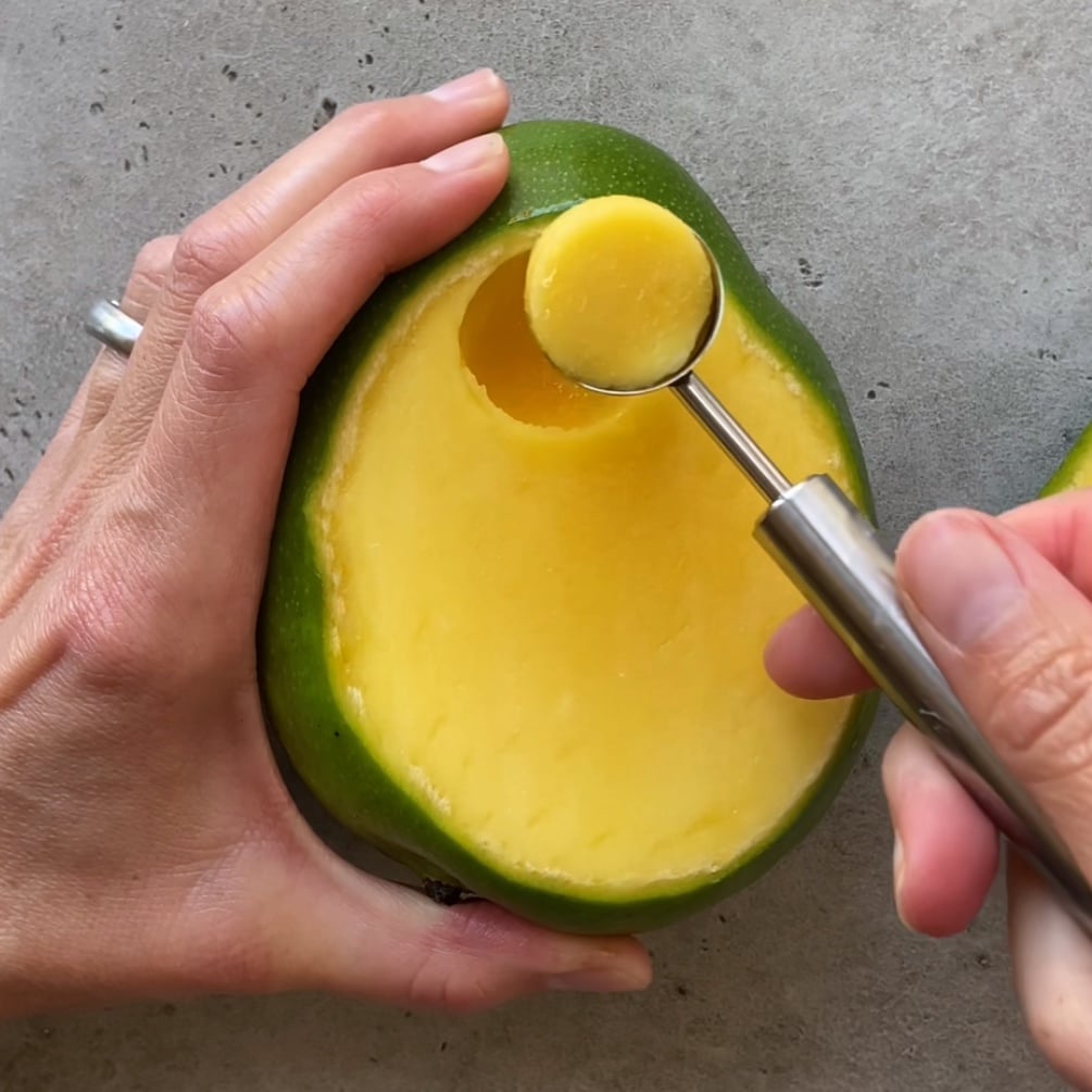 A person uses a melon baller to scoop a round piece from the flesh of a halved mango on a gray surface.