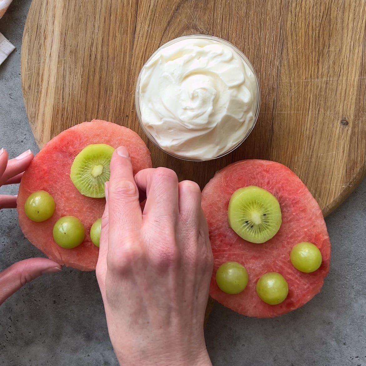 Two hands arrange kiwi slices and green grapes on watermelon pieces for an Easter Fruit Tray.