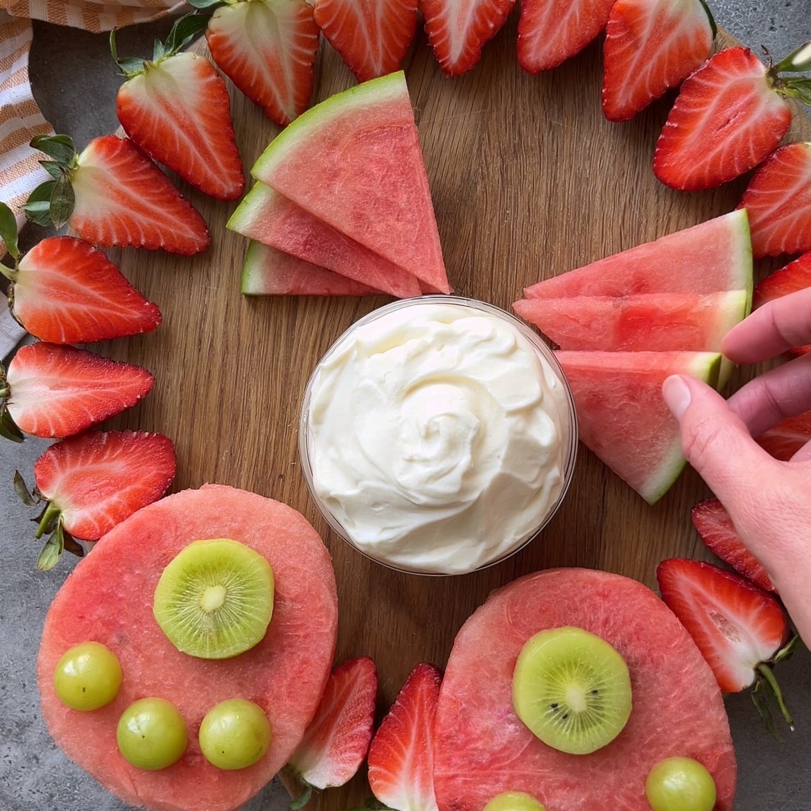 A hand places a watermelon slice on a wooden board, arranged with sliced strawberries, watermelon, and a bowl of dip.
