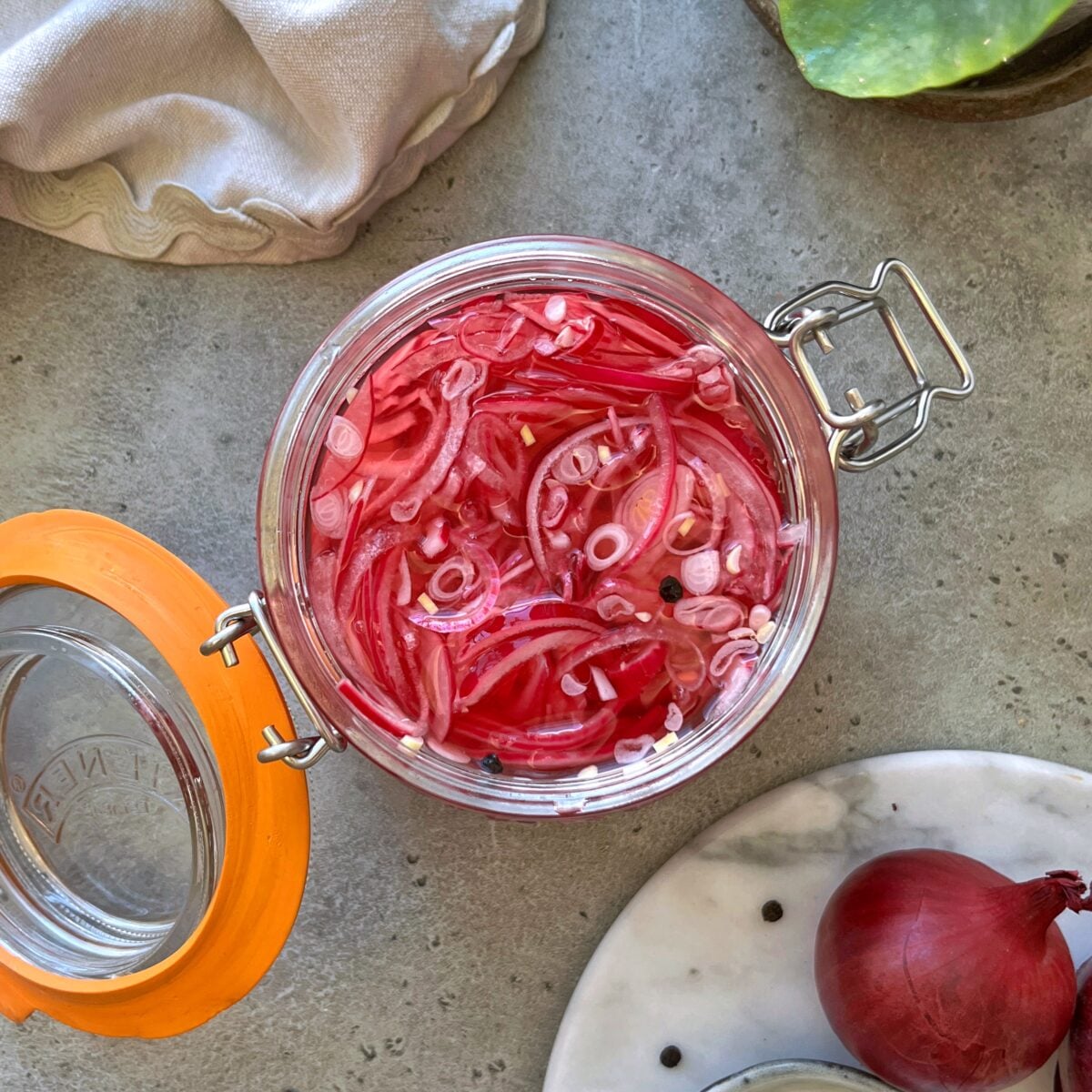 An open glass jar of Easy Pickled Red Onions sits on a gray countertop beside a whole red onion.