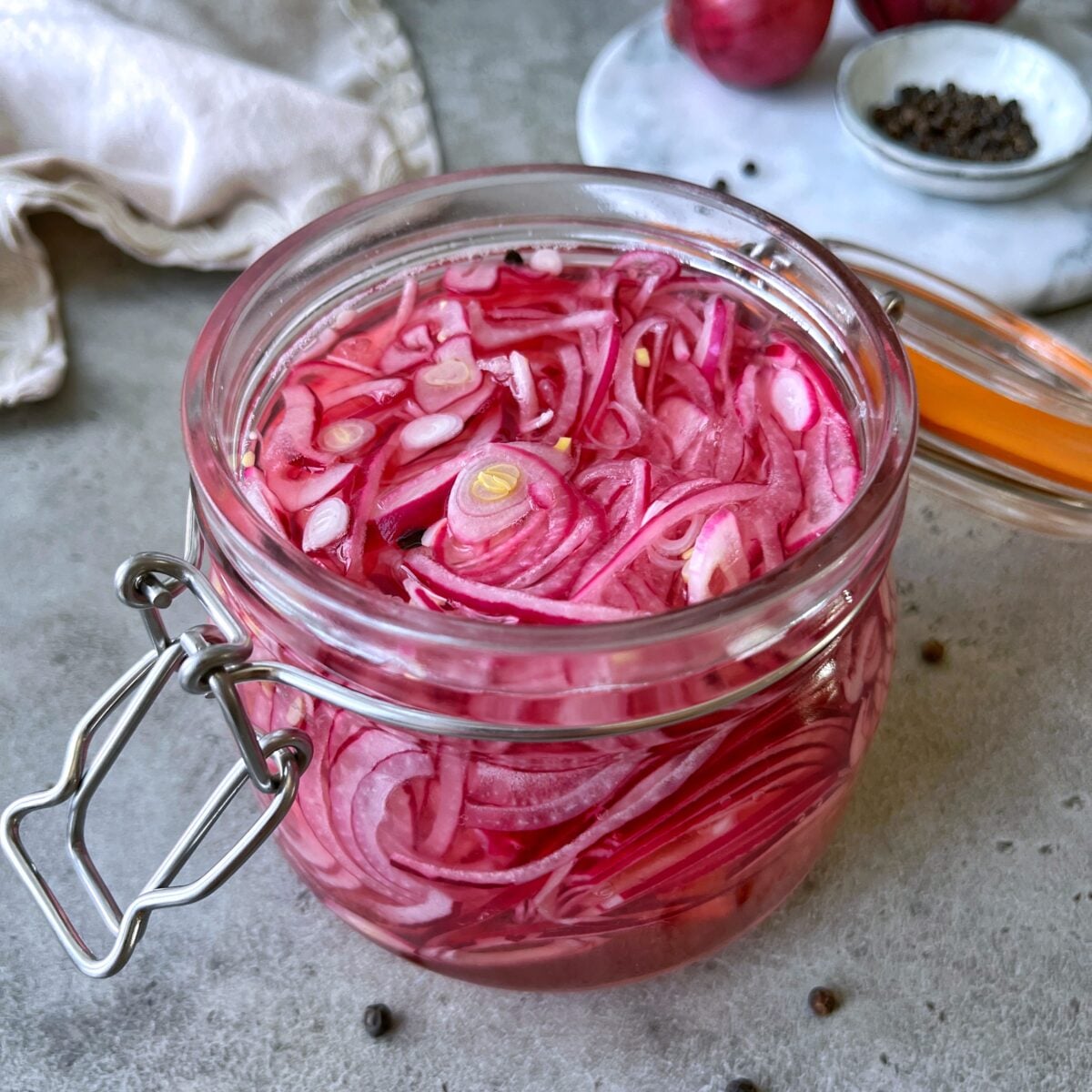 A glass jar filled with Easy Pickled Red Onions in tangy pickling liquid, placed on a gray surface with scattered peppercorns and a cloth nearby.