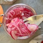 A fork lifts thinly sliced Easy Pickled Red Onions from a glass jar filled with vibrant pickled onions, set on a gray surface.