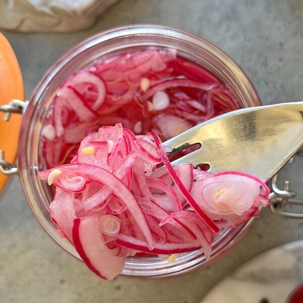 A fork lifts thinly sliced Easy Pickled Red Onions from a glass jar filled with vibrant pickled onions, set on a gray surface.