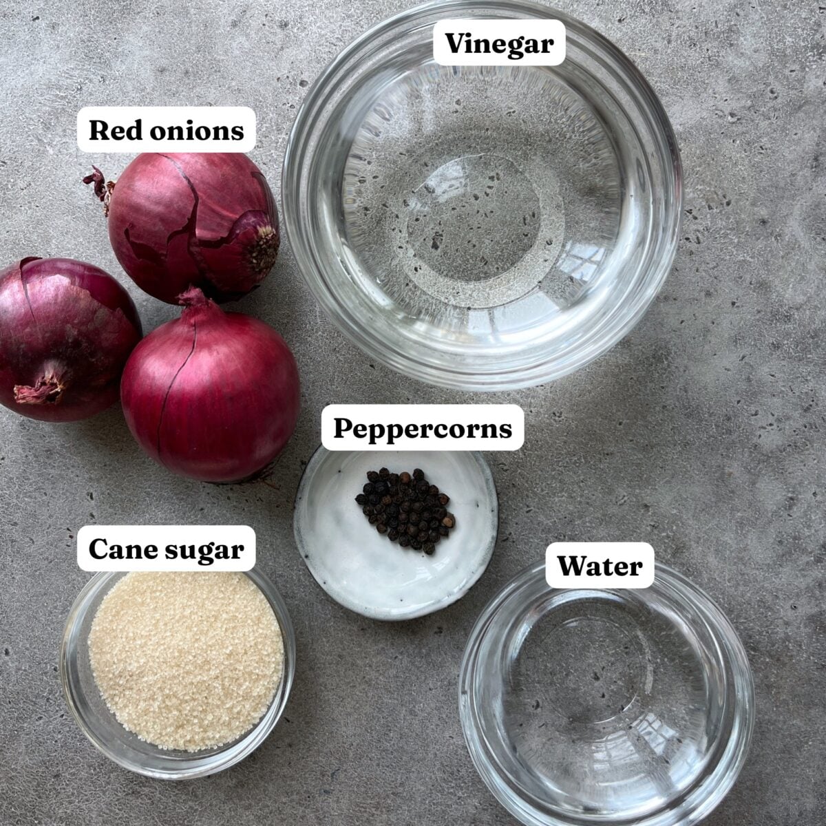 Overhead view of red onions, cane sugar, peppercorns, vinegar, and water in separate bowls on a gray surface, each ingredient labeled.