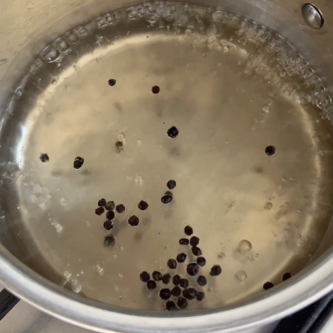 A stainless steel pot of water, vinegar, and sugar is boiling on a stove with whole black peppercorns floating on the surface.