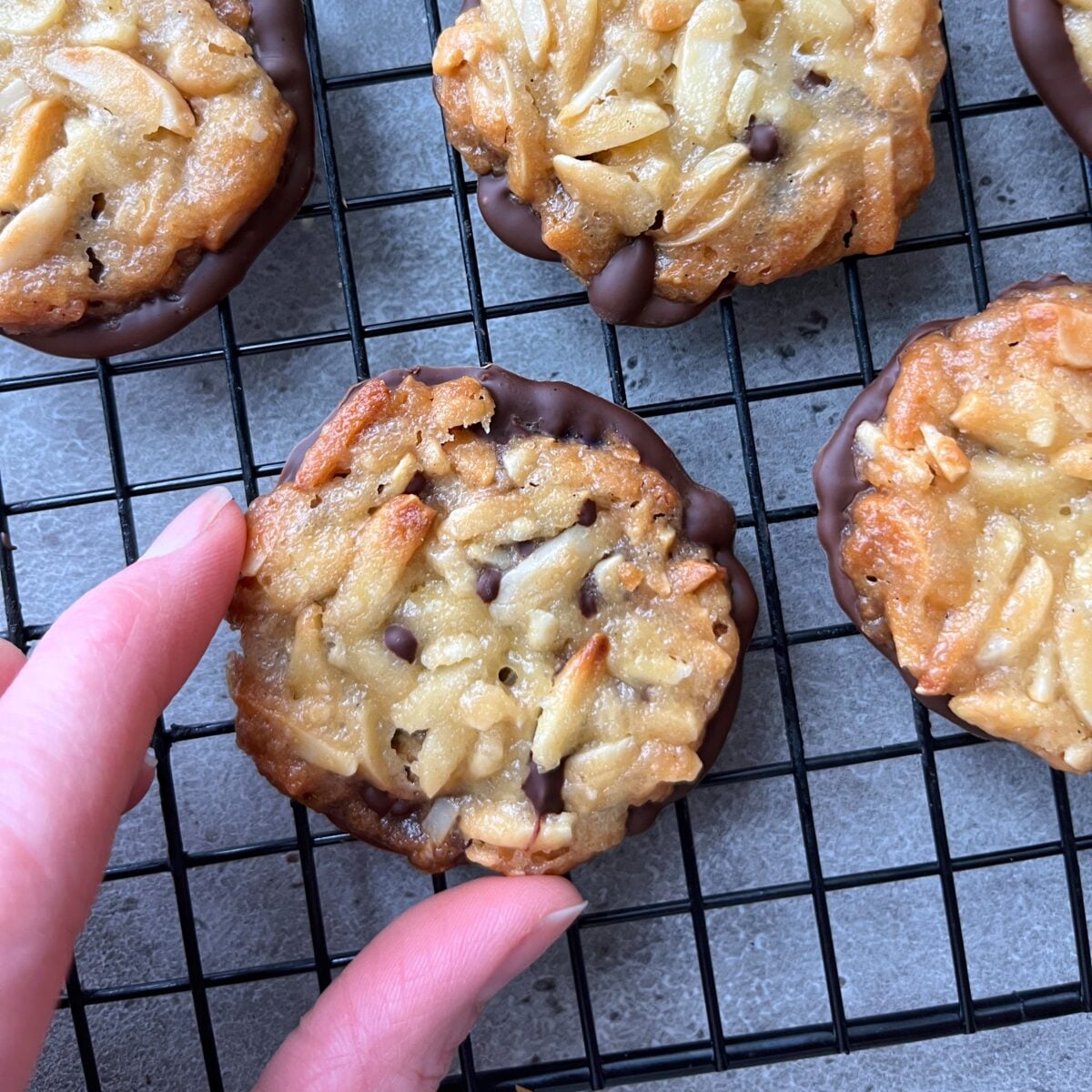 Hand reaching for a Florentine cookie with sliced almonds and chocolate resting on a black cooling rack alongside other Florentines.