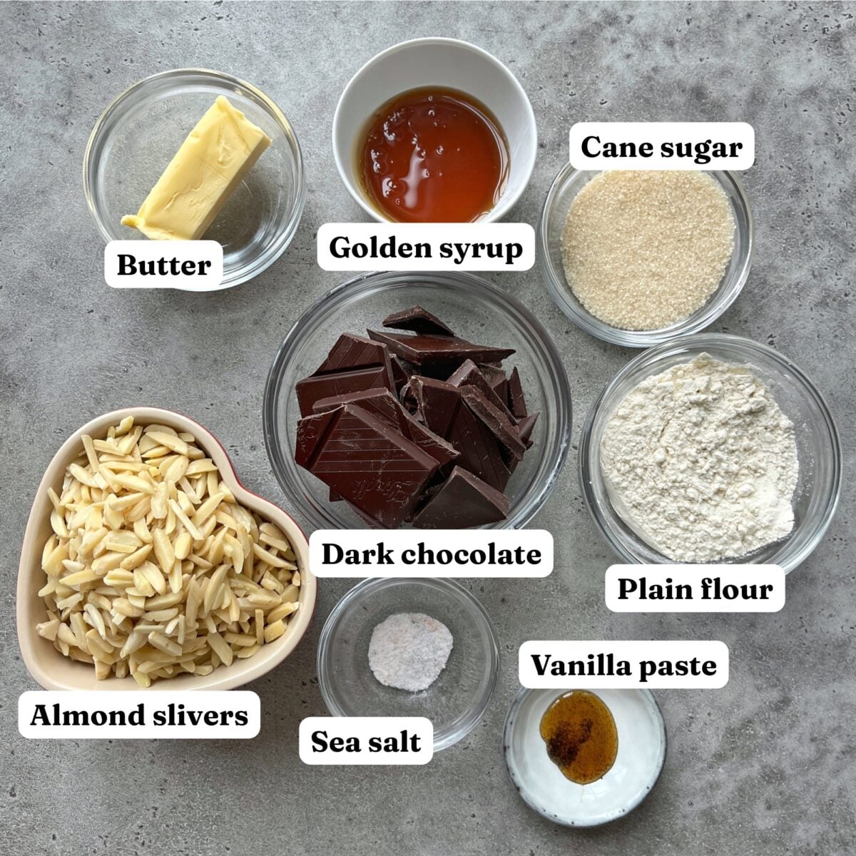 Top-down view of labeled Florentines ingredients in bowls: butter, golden syrup, cane sugar, plain flour, vanilla paste, sea salt, almond slivers, and dark chocolate on a grey surface.