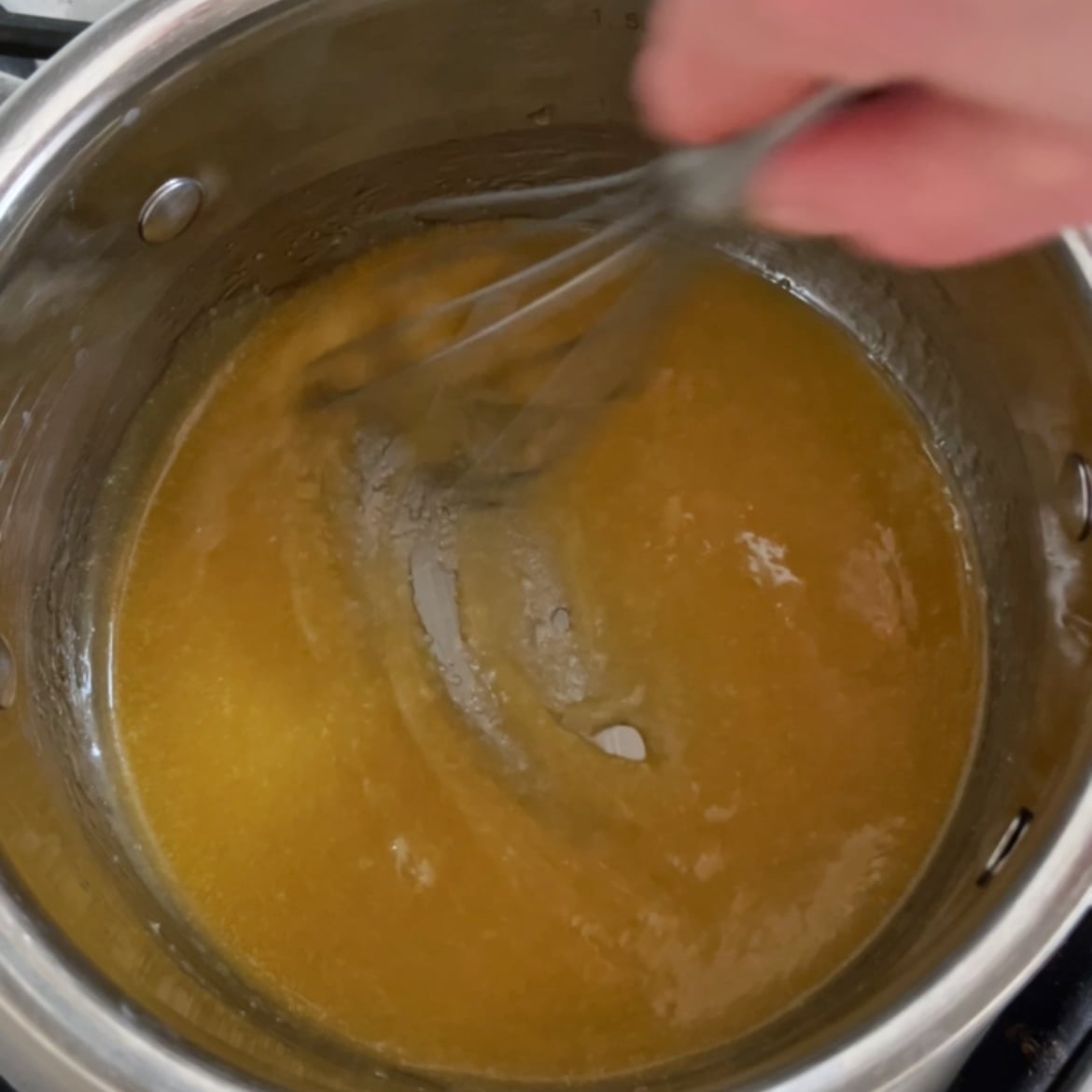 A hand uses a whisk to stir a caramel mixture in a stainless steel pot on a stovetop.