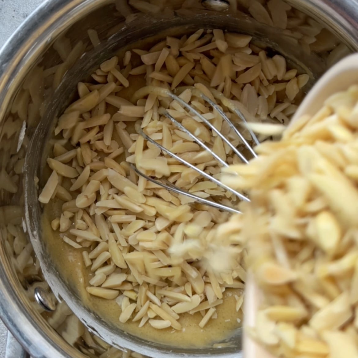 Slivered almonds are being added to a saucepan containing a whisk and a caramel.