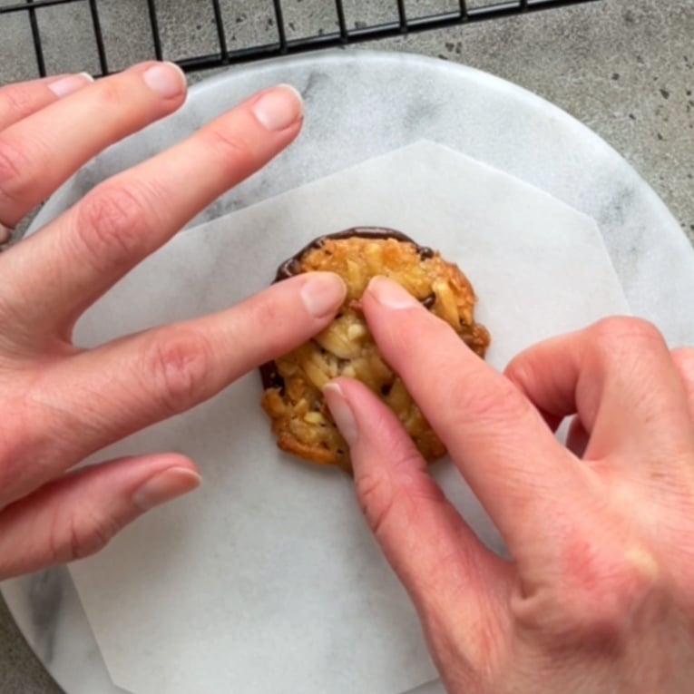 Two hands pressing a round, nut-filled Florentine cookie onto a blob of dark chocolate.