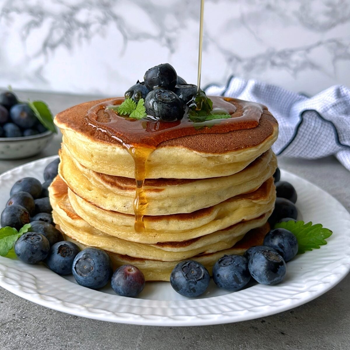 A stack of Greek Yogurt Pancakes topped with blueberries and mint, with syrup being poured over them, served on a white plate with more blueberries around the base.