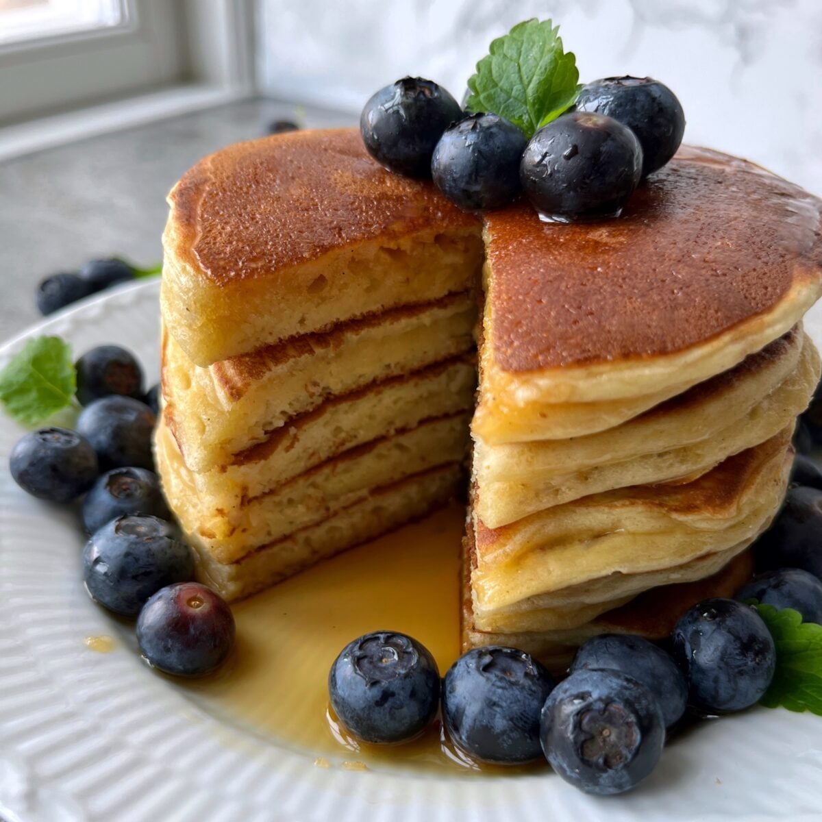 A close up of a stack of Greek Yogurt Pancakes with a wedge cut out, topped with blueberries and syrup, served on a white plate with extra blueberries scattered around.