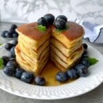A stack of Greek Yogurt Pancakes with a wedge cut out, topped with blueberries and syrup, served on a white plate with extra blueberries and a mint garnish.