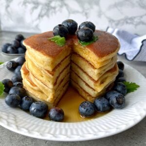 A stack of Greek Yogurt Pancakes with a wedge cut out, topped with blueberries and syrup, served on a white plate with extra blueberries and a mint garnish.