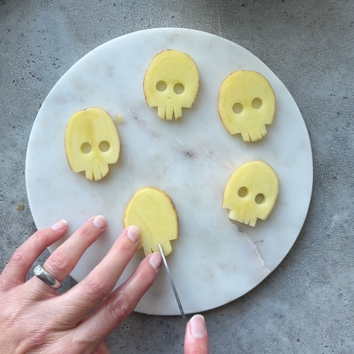 Four thin potato slices cut into skull shapes are arranged on a round marble board. A hand is carving details into another slice.