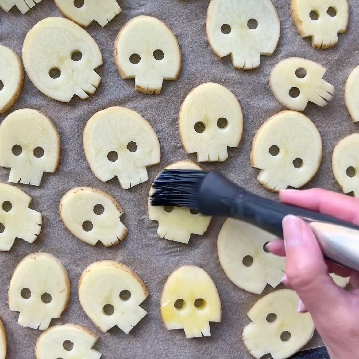 Potato slices cut into skull shapes are arranged on a baking sheet. A hand is brushing oil onto the slices with a basting brush.