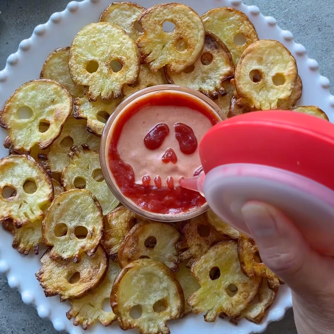 A plate of Halloween Fries shaped like skulls surrounds a bowl of dip decorated with a skull design in ketchup.