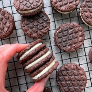 A hand holds three Homemade Oreos with cream filling over a cooling rack filled with more cookies.