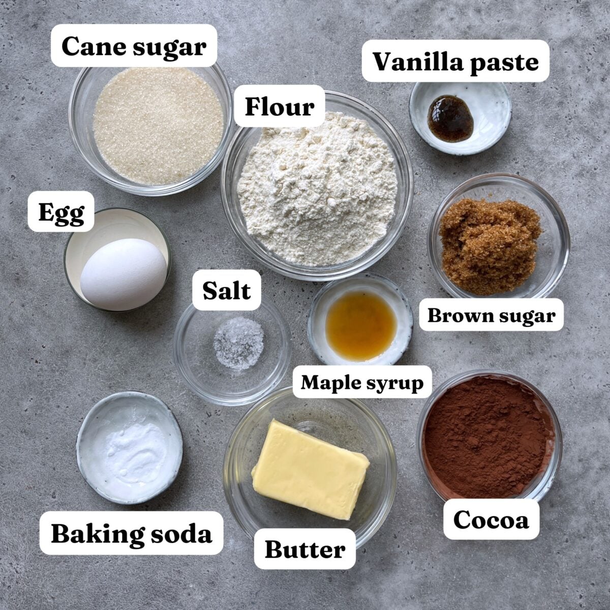 Top-down view of baking ingredients in bowls, including cane sugar, egg, baking soda, salt, butter, maple syrup, flour, vanilla paste, brown sugar, and cocoa.