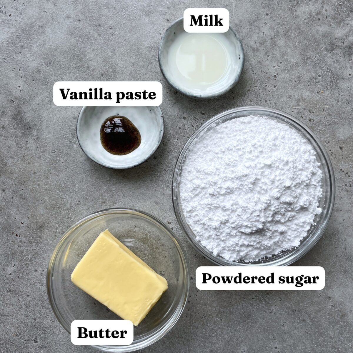 Four labeled ingredients for frosting—milk, vanilla paste, powdered sugar, and butter—are arranged in small bowls on a gray surface, perfect for pairing with crushed Oreo Cookies.