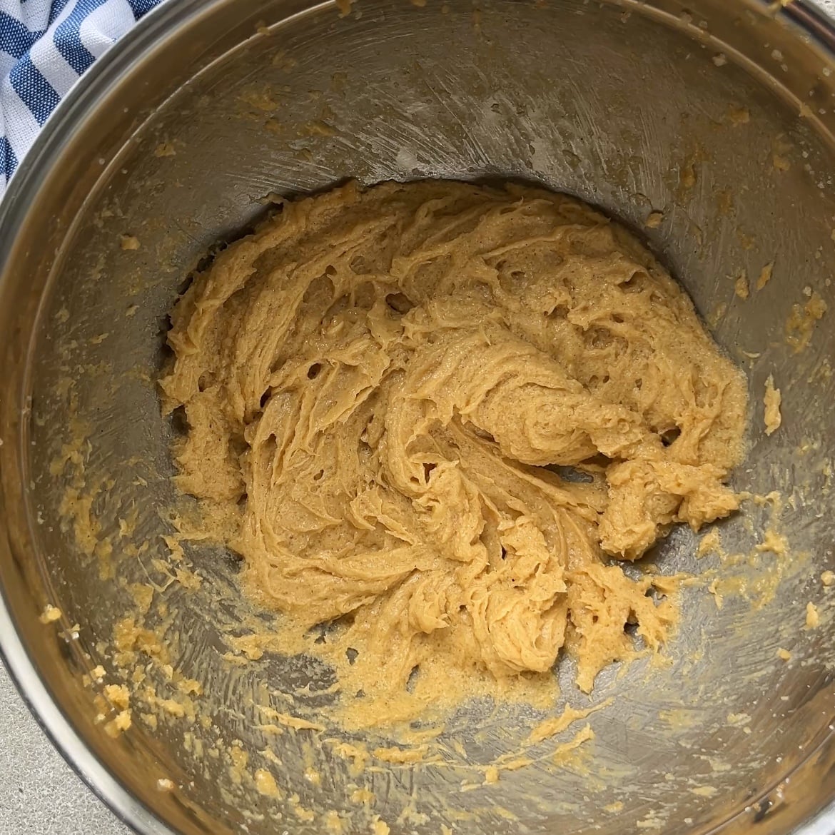A metal mixing bowl containing creamed cookie dough mixture sitting on a countertop with a blue and white striped cloth partially visible.