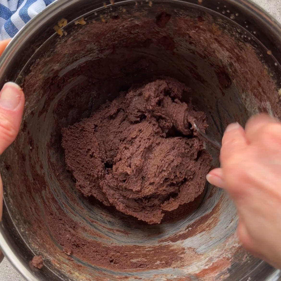 A person mixing thick cookie batter in a metal bowl with a spoon.