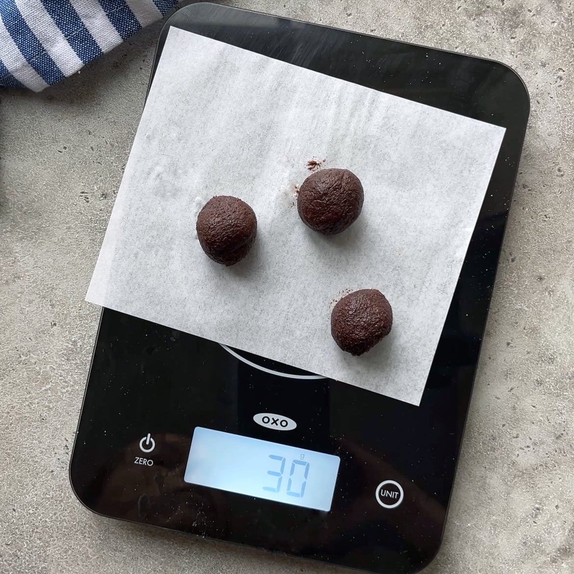 Three small chocolate dough balls are placed on parchment paper atop a digital kitchen scale, displaying a weight of 30 grams.