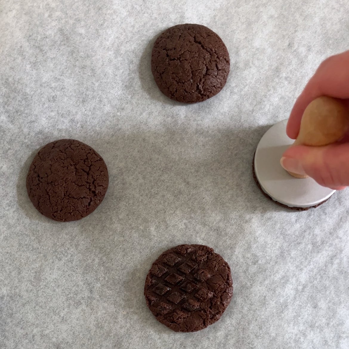 A hand uses a cookie stamp on a chocolate cookie, laid on parchment paper with three other chocolate cookies, one displaying a patterned surface.