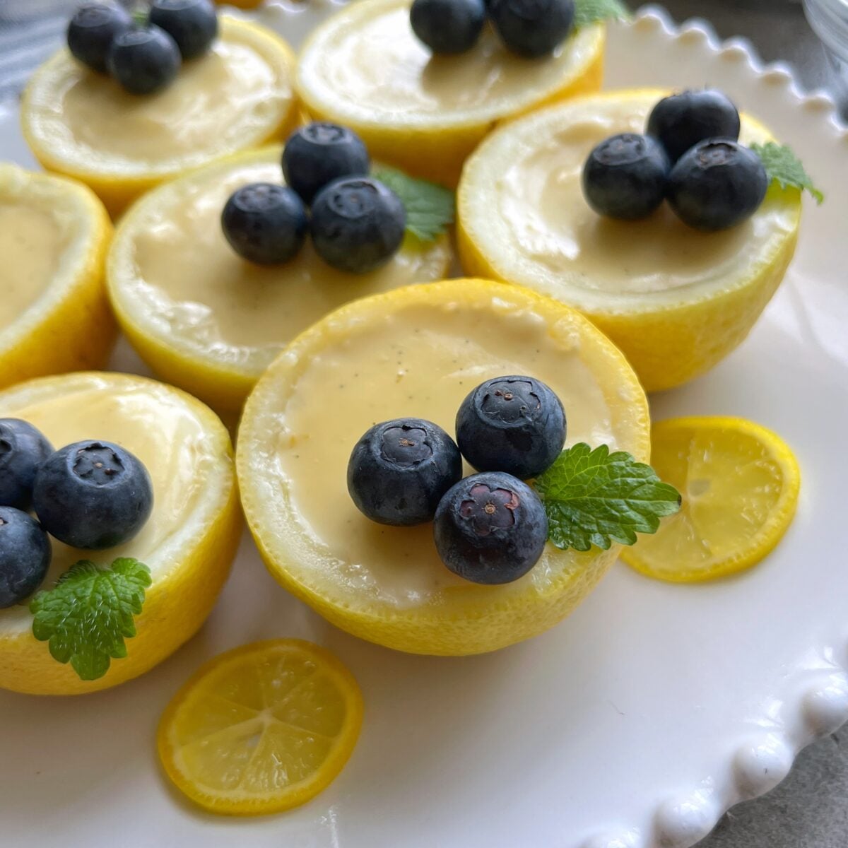 Lemon Posset halves filled with creamy custard, topped with blueberries and a mint leaf, arranged on a white plate with fresh lemon slices.