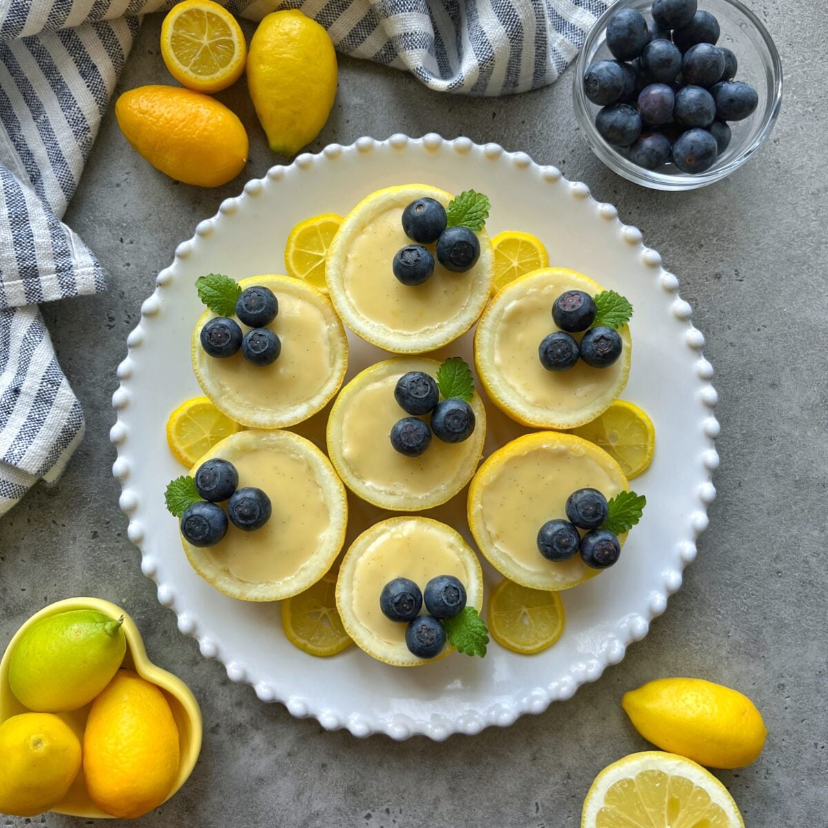 A white plate holds seven mini lemon posset tarts topped with blueberries and mint leaves, surrounded by lemon slices, whole lemons, and a bowl of blueberries.