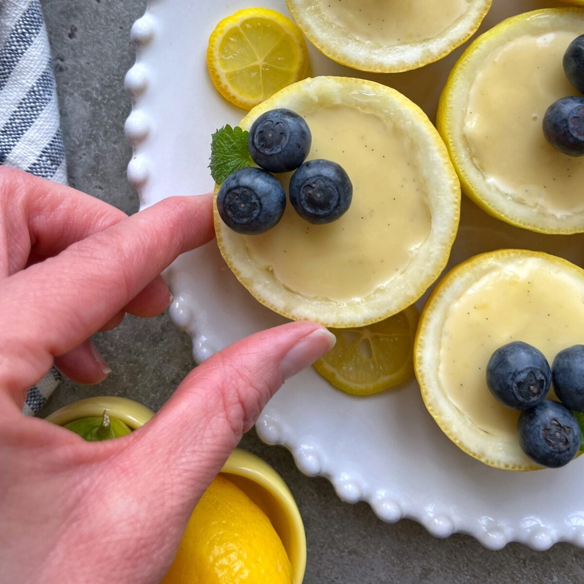 A hand picks up a lemon half filled with creamy Lemon Posset and topped with three blueberries from a white plate with similar desserts.