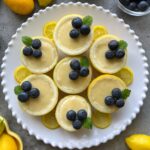 Seven mini lemon possets topped with blueberries and mint leaves are arranged on a white plate with lemon slices.
