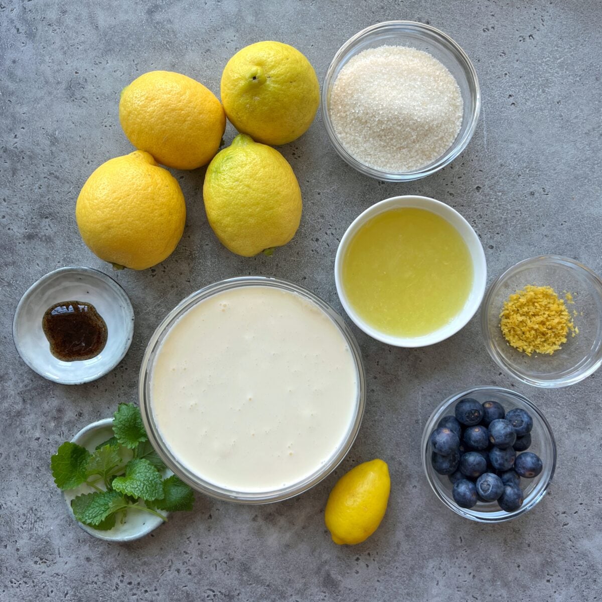 Assorted ingredients on a gray surface, including lemons, sugar, lemon juice, lemon zest, blueberries, fresh mint, heavy cream, and a small bowl of vanilla paste.
