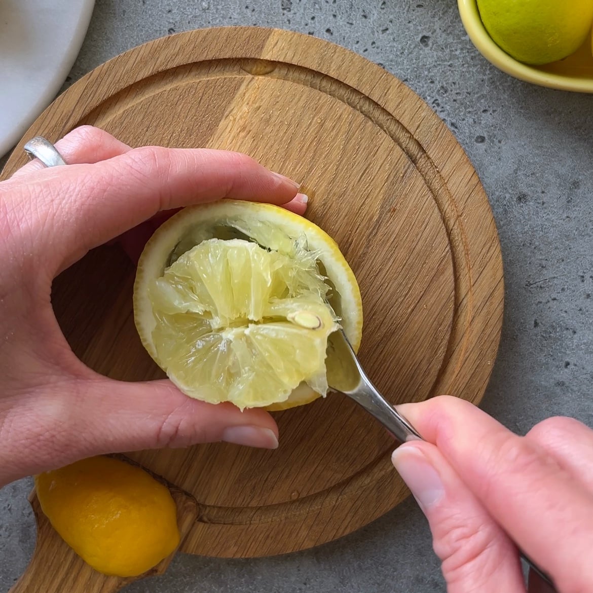 A hand uses a spoon to remove the flesh from a halved lemon on a round wooden cutting board.