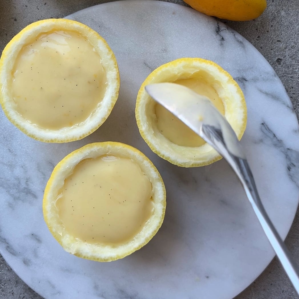 Three lemon halves are being filled with creamy Lemon Posset.