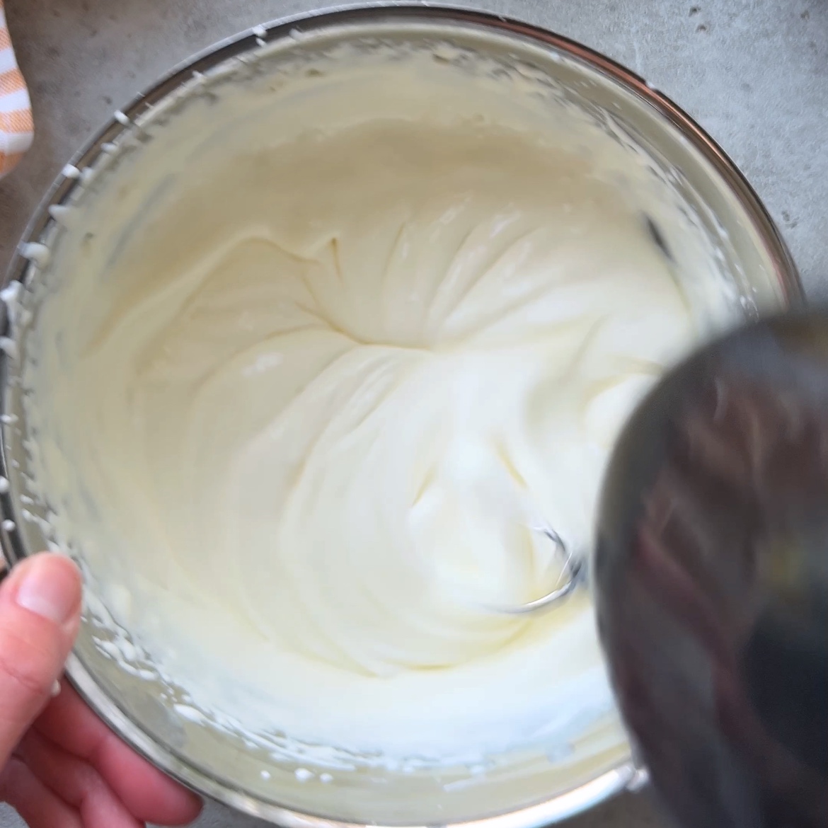 A hand holds a mixing bowl filled with creamy mascarpone and heavy cream whipped until soft peaks form.