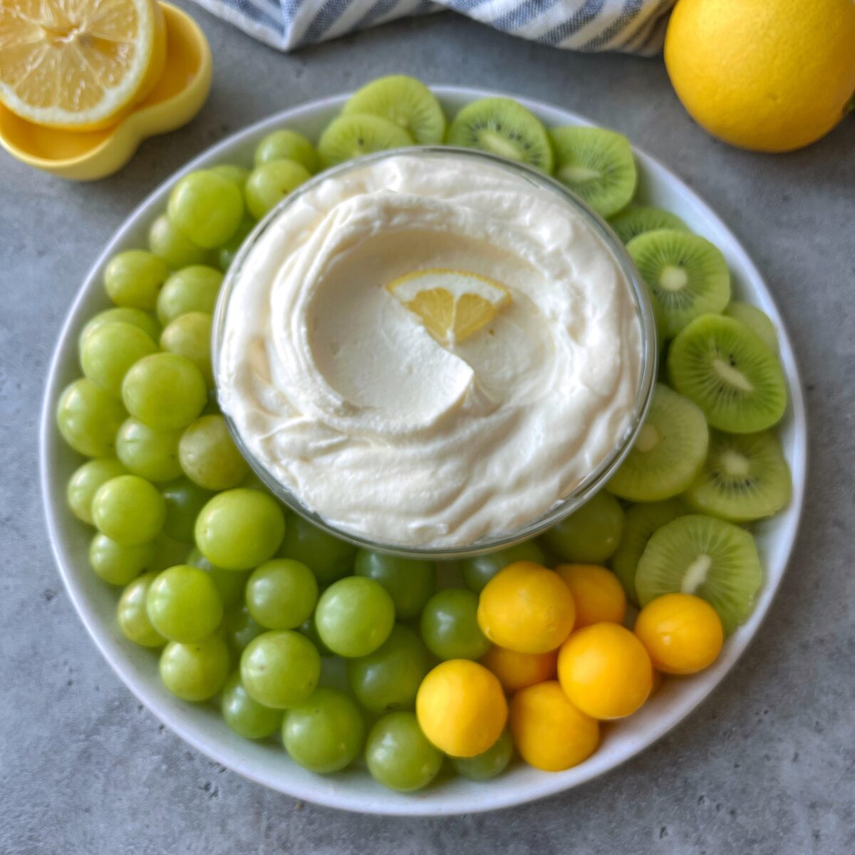 Plate with green grapes, sliced green kiwis, mango balls, and a bowl of creamy lemon dip garnished with a lemon wedge in the center.