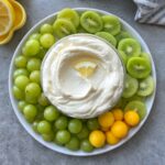 A plate with green grapes, sliced green kiwifruit, mango balls, and a bowl of creamy dip garnished with a lemon wedge in the center.