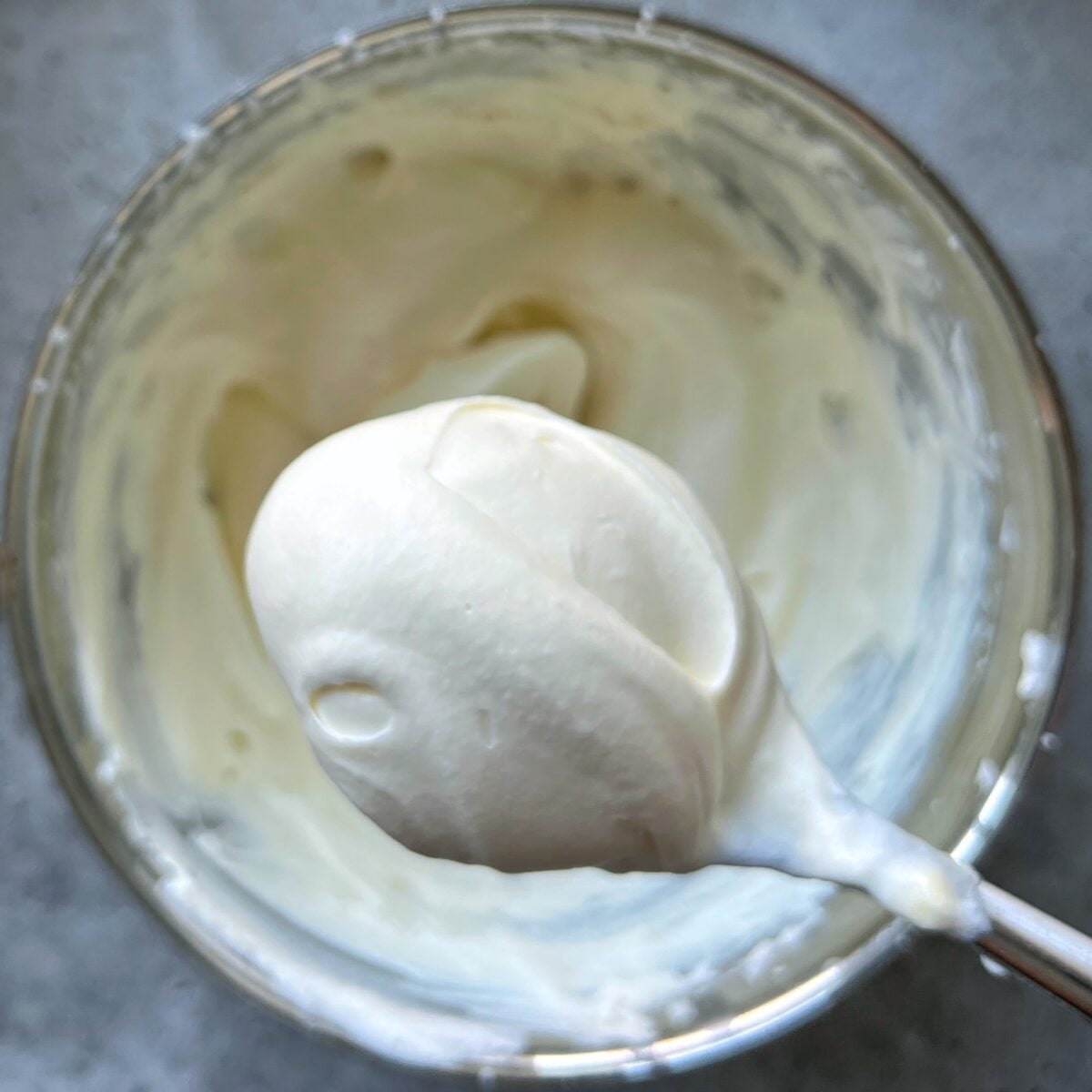 A spoonful of thick, creamy white Mascarpone Fruit Dip is held above a bowl filled with the same luscious mixture on a gray surface.