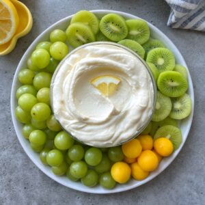 A plate with green grapes, sliced green kiwifruit, mango balls, and a bowl of creamy dip garnished with a lemon wedge in the center.