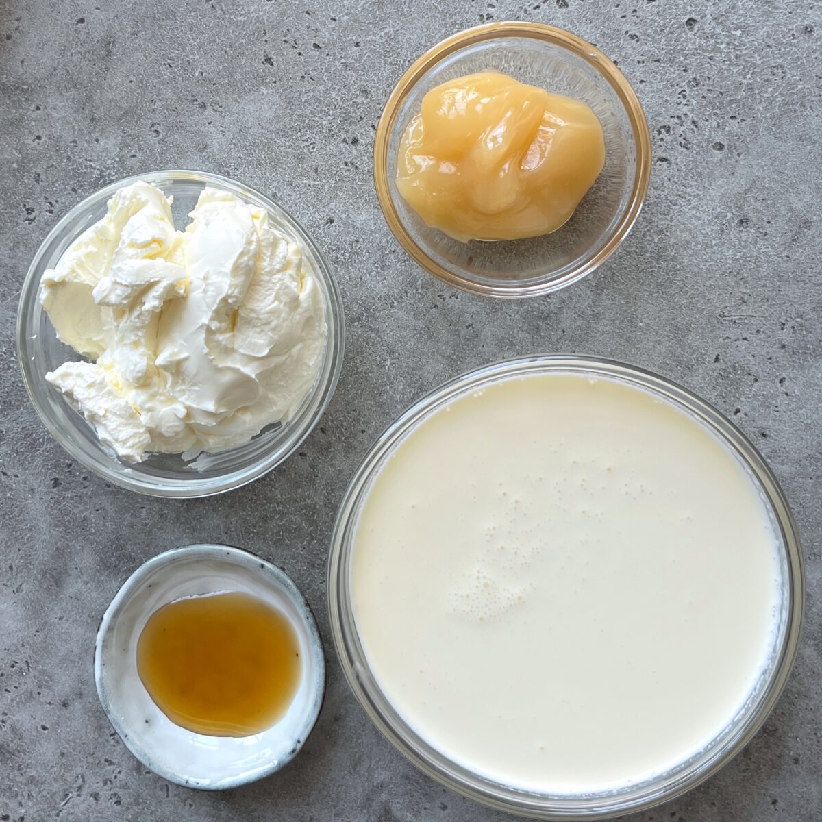 Four bowls on a gray surface contain heavy cream, mascarpone cheese, maple syrup, and lemon curd.