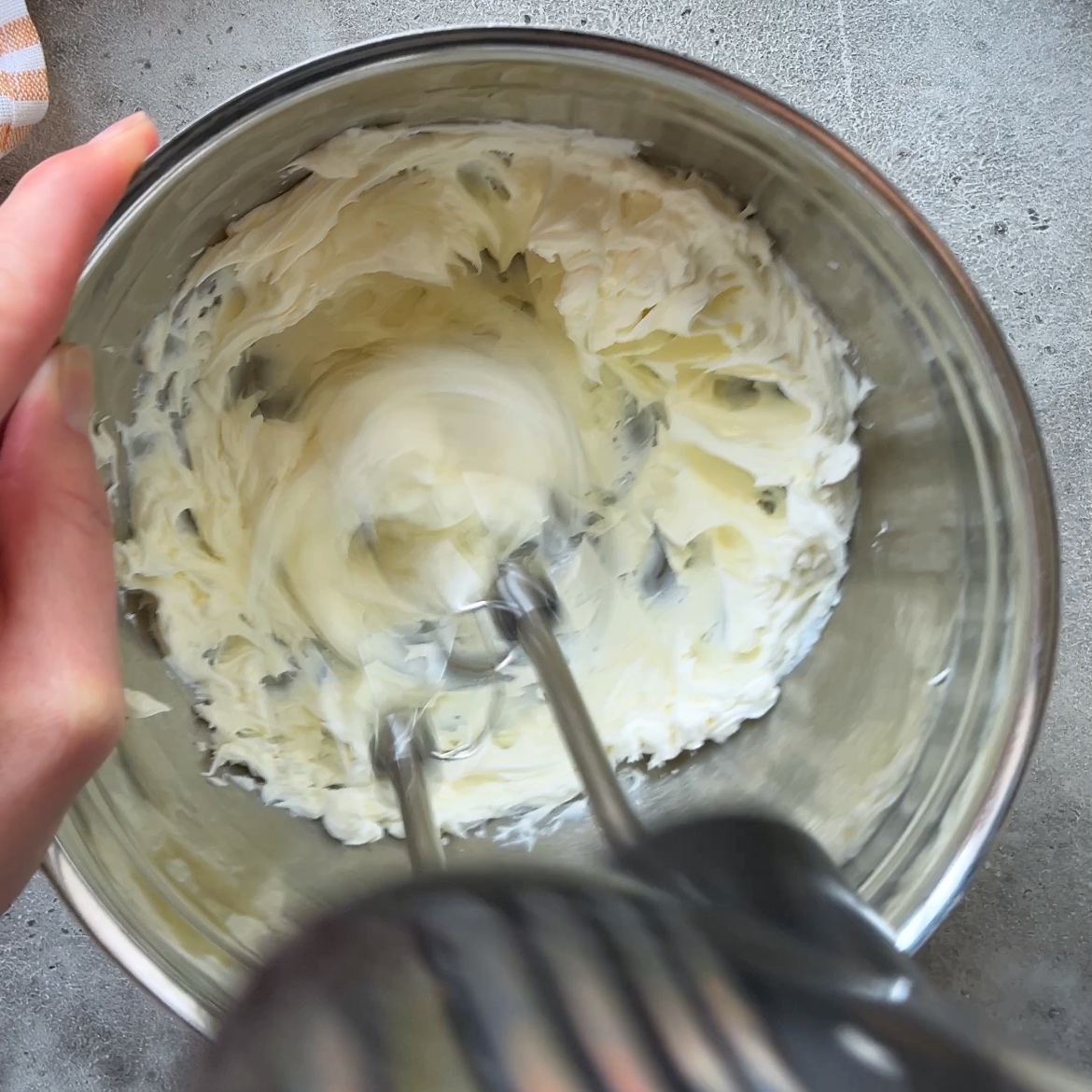 A hand holds an electric mixer blending mascarpone for 1 minute.