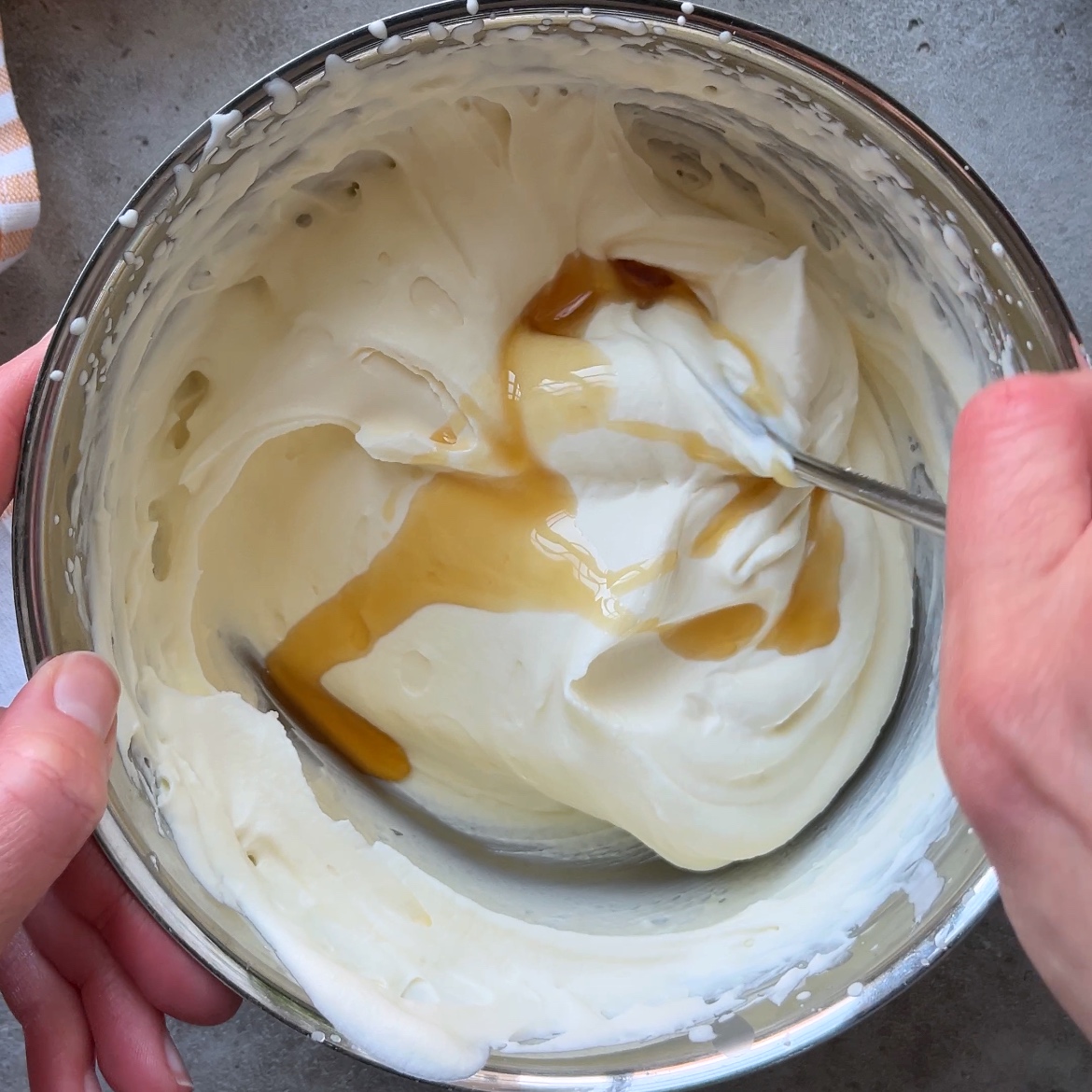 A person stirs a metal bowl of whipped mascarpone cream with a spoon, while a stream of maple syrup is being added.