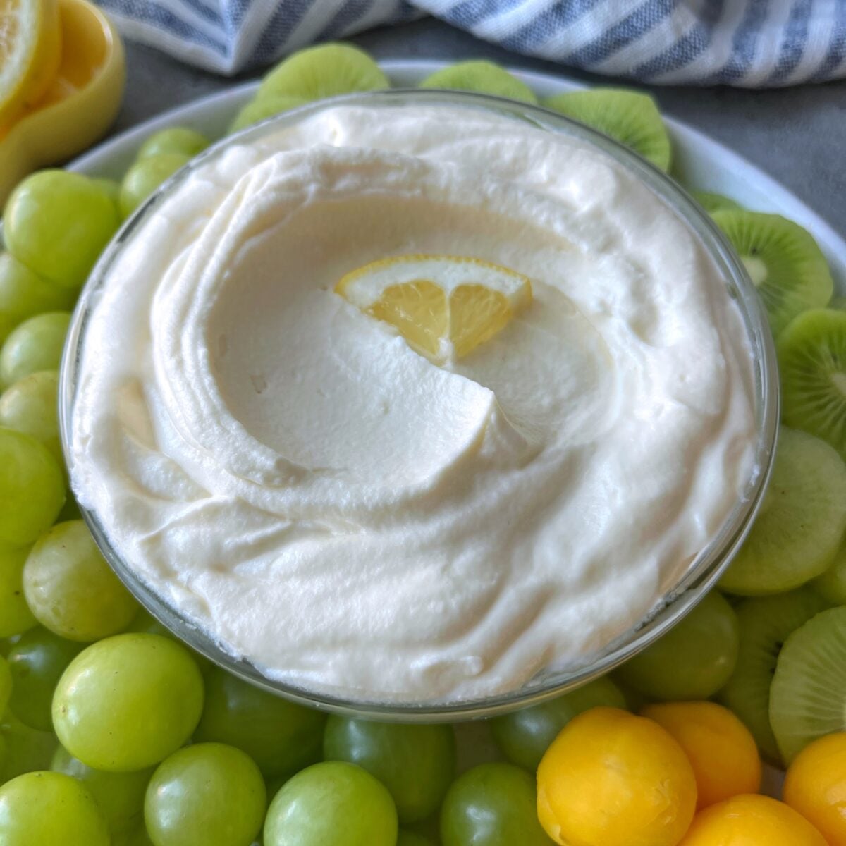 A bowl of creamy Mascarpone Fruit Dip topped with a lemon slice, surrounded by green grapes, mango balls, and sliced kiwi is ready to be served.