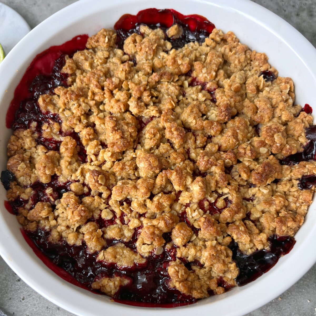 A baked fruit crumble dessert in a white round dish, topped with a golden-brown oat topping.
