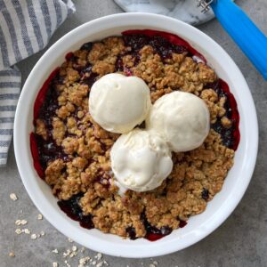 A round dish of berry crumble topped with three scoops of vanilla ice cream sits on a gray surface next to a striped cloth.